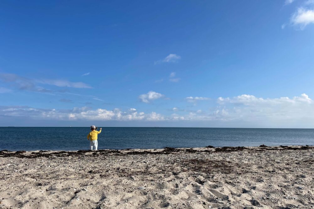 Blauer Himmel, blaues Meer und Strand. Kind mit gelbem Pullover steht am Wasser.