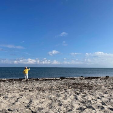 Blauer Himmel, blaues Meer und Strand. Kind mit gelbem Pullover steht am Wasser.