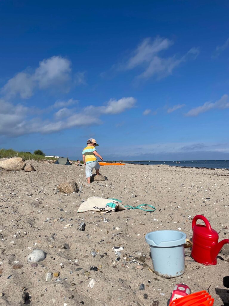 Leicht steiniger Strand mit blauem Eimer und roter Gieskann. Kleinkind klettert auf kniehohen Stein. 